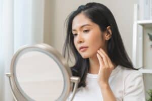 Woman looks in the mirror with hands on her face, looking at her skin, considering cosmetic dermatology.